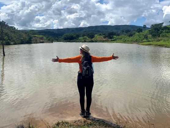 mulher caminhante na beira de uma lago com mochila nas costas e braços abertos horizontalmente aprecia a paisagem composta por vegetação verde ao redor do lago montanhas ao fundo e céu azul coberto por nuvens